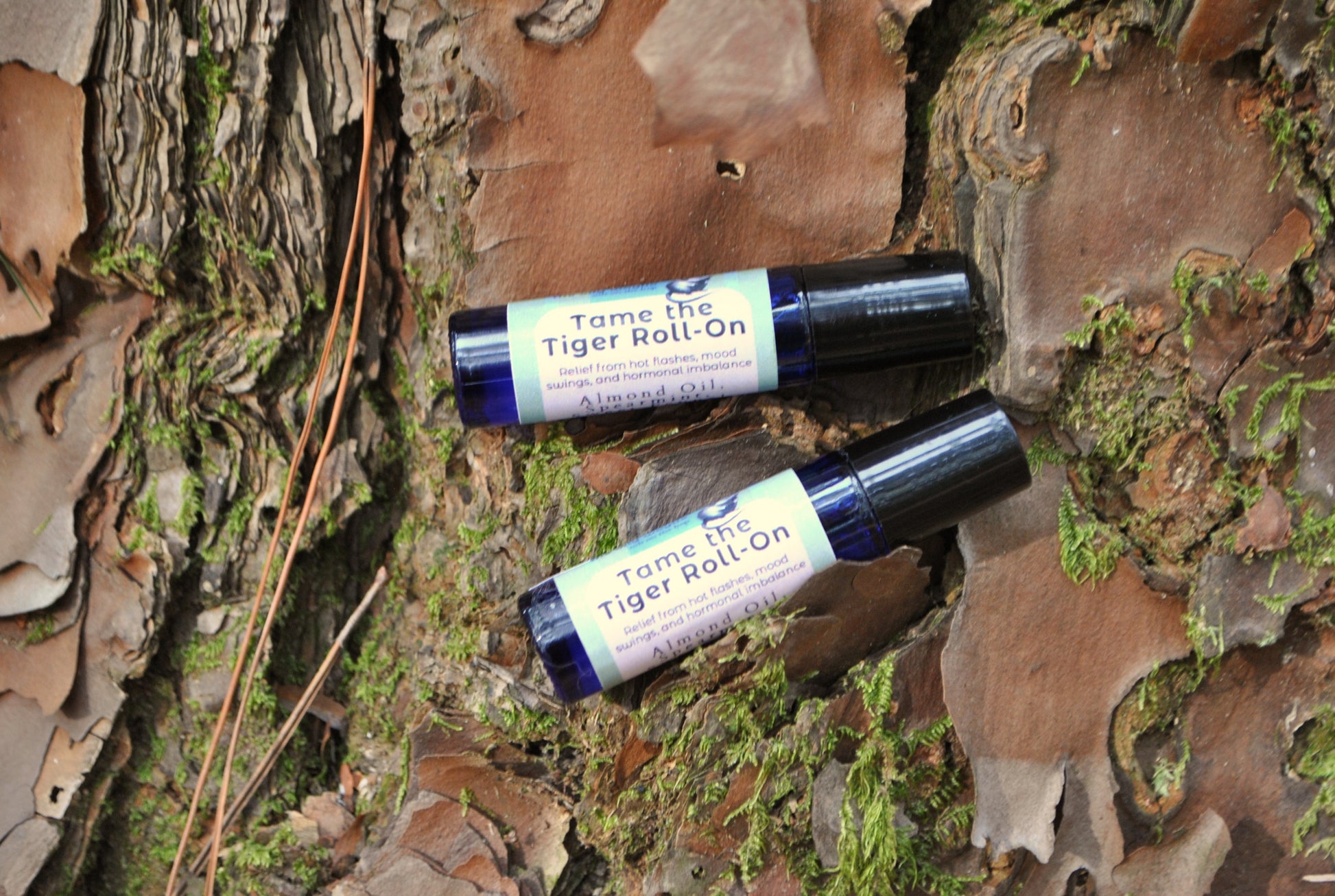 Two bottles of 'Tame the Tiger Roll-On' on a natural background with tree bark and moss.