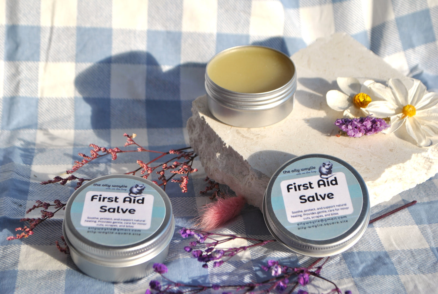 Three tins of 'First Aid Salve' on a checkered fabric background with flowers and leaves.