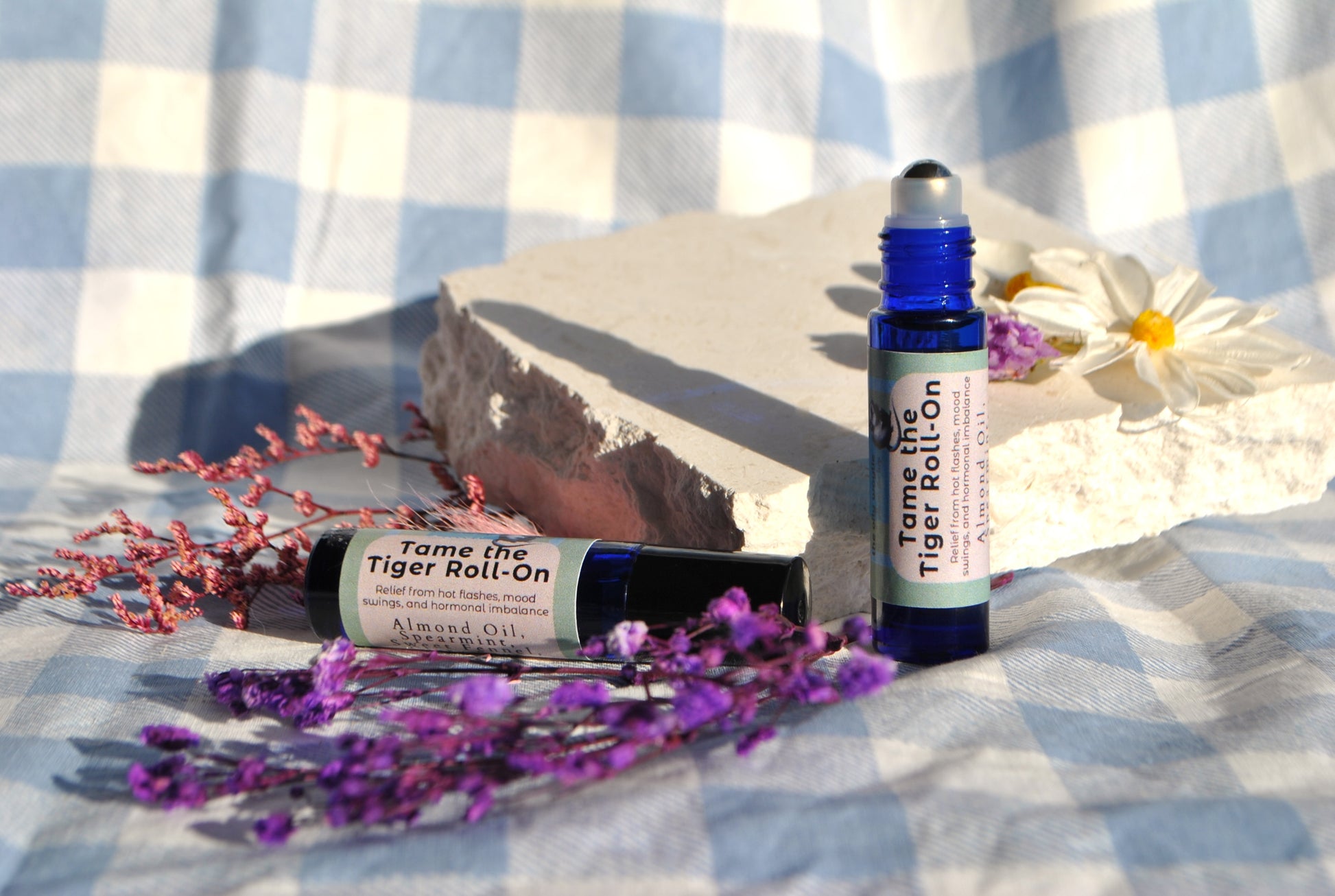 Two roller bottles labeled 'Tame the Tiger Roll-On' on a checkered fabric background with flowers.