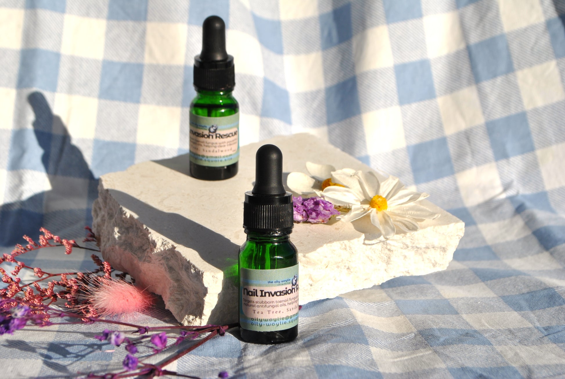Two bottles of 'Nail Invasion' on a stone surface with flowers and a checkered cloth background