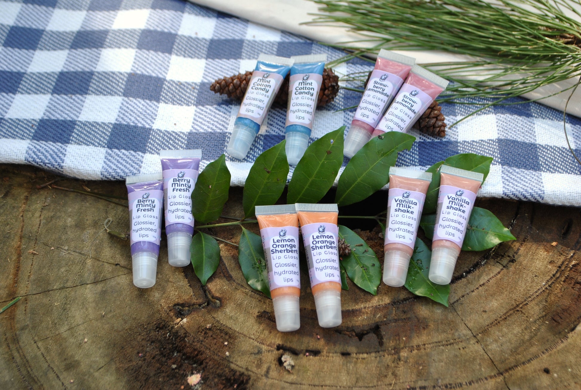 Assorted lip gloss tubes on a wooden surface with leaves and a checkered cloth in the background