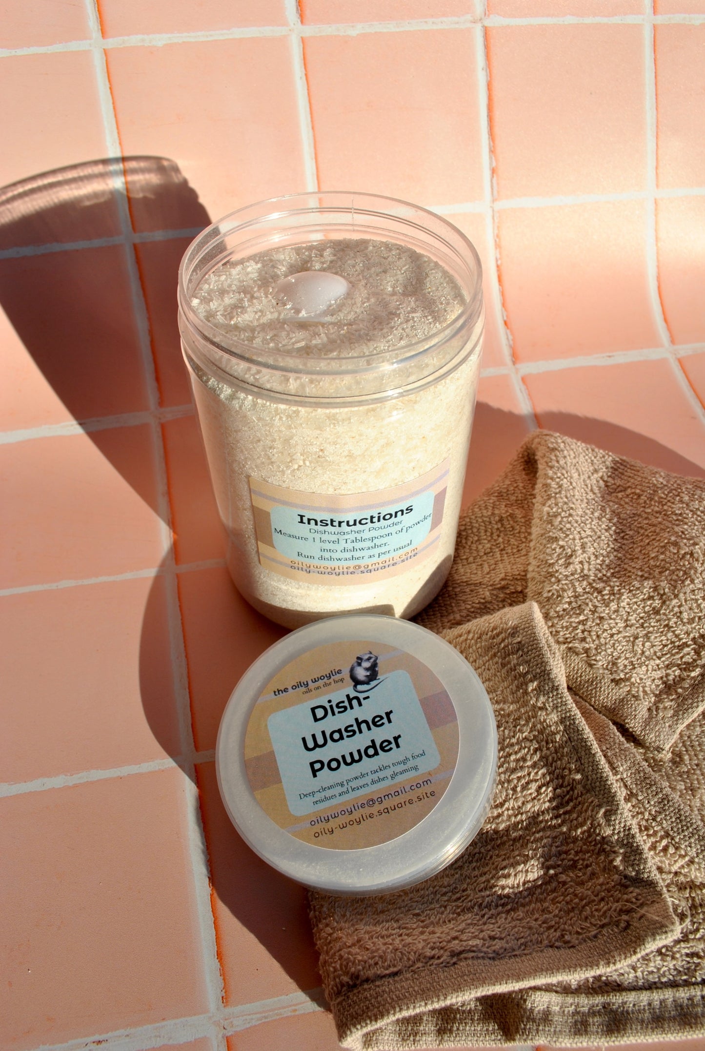 Two containers of dishwasher powder on a tiled surface with a brown towel.