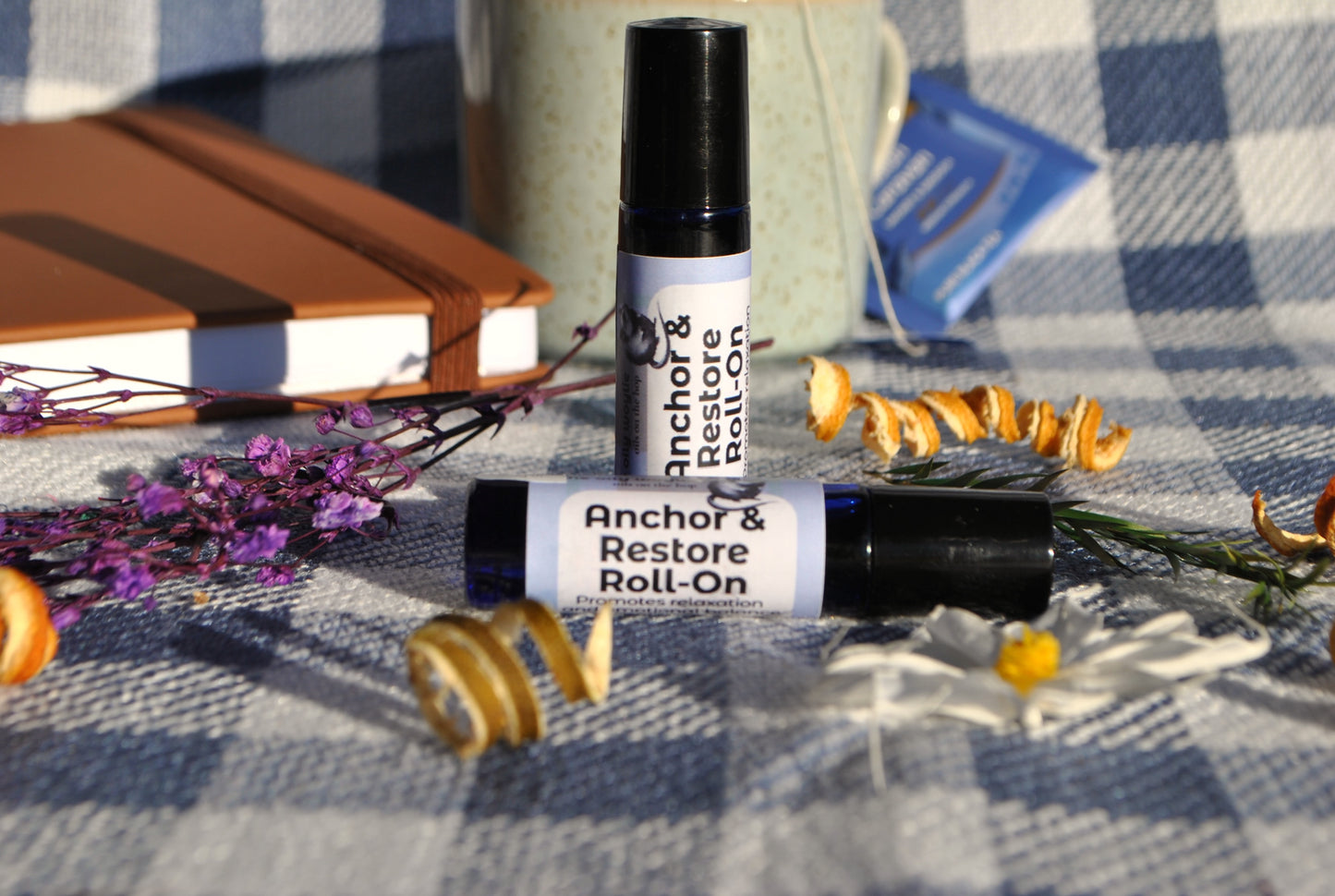 Anchor & Restore Roll essential oil roller blend, grounding and restorative support on a checkered tablecloth with flowers and a notebook.