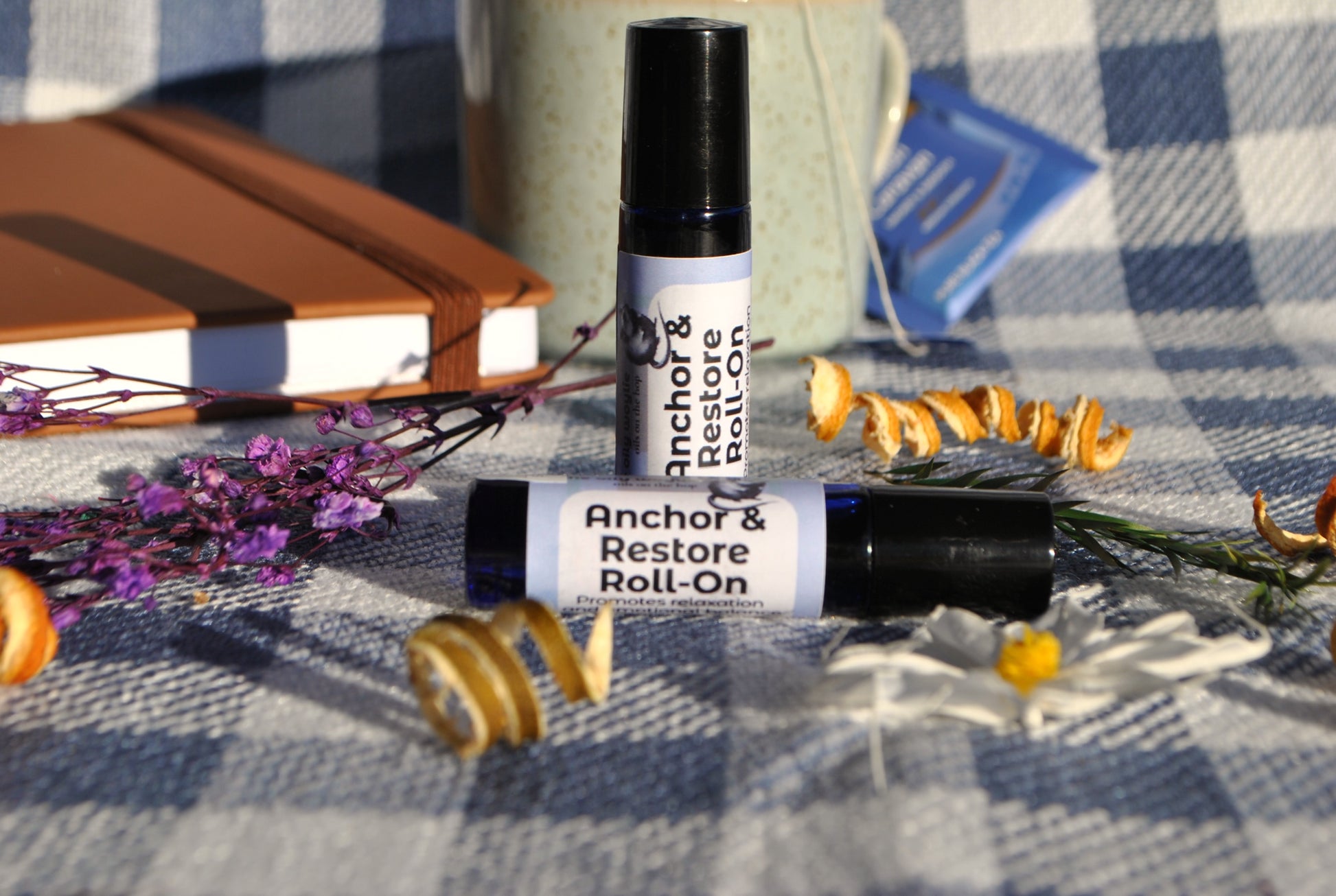 Anchor & Restore Roll essential oil roller blend, grounding and restorative support on a checkered tablecloth with flowers and a notebook.