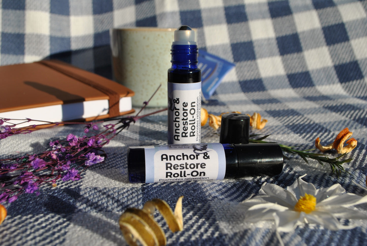 Anchor & Restore Roll essential oil roller blend, grounding and restorative support Two Roll-On bottles on a checkered fabric background with flowers and a notebook.