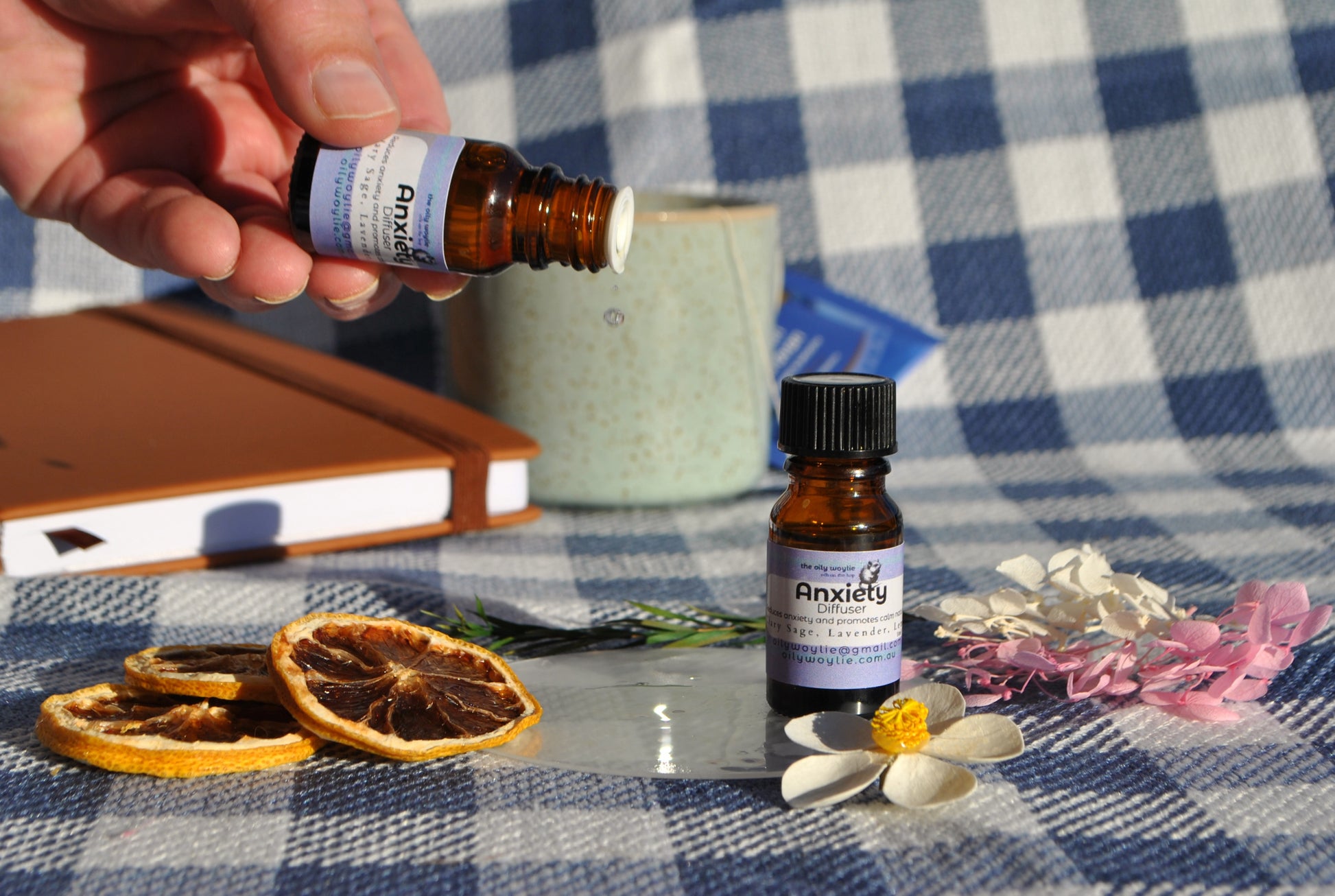 Anxiety Diffuser essential oil blend bottle, natural support for calm and relaxation on a checkered tablecloth with dried oranges and flowers. Dripping