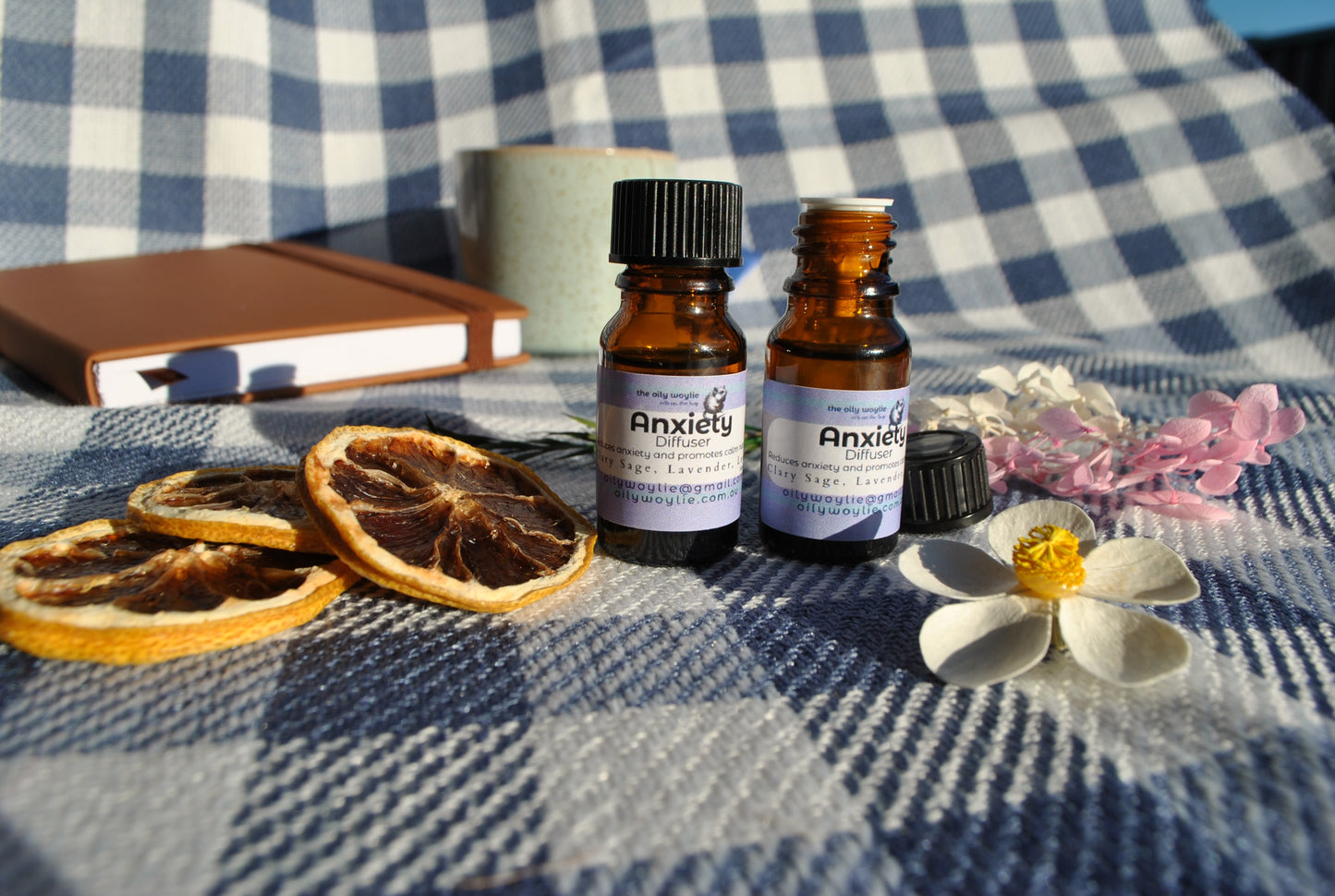 Anxiety Diffuser essential oil blend bottle, natural support for calm and relaxation on a checkered fabric surface with dried oranges and flowers. Lid off