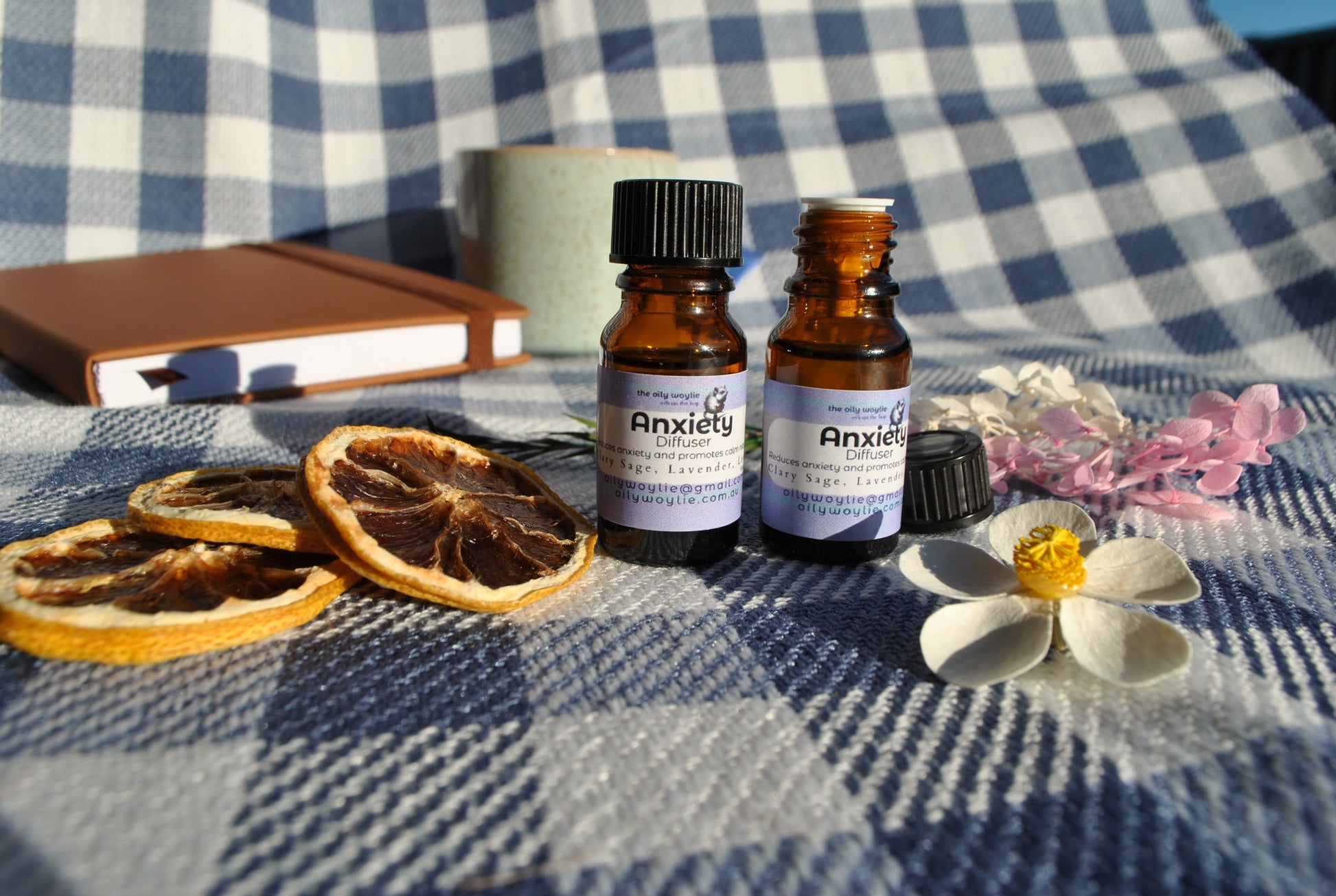 Anxiety Diffuser essential oil blend bottle, natural support for calm and relaxation on a checkered fabric surface with dried oranges and flowers. Lid off