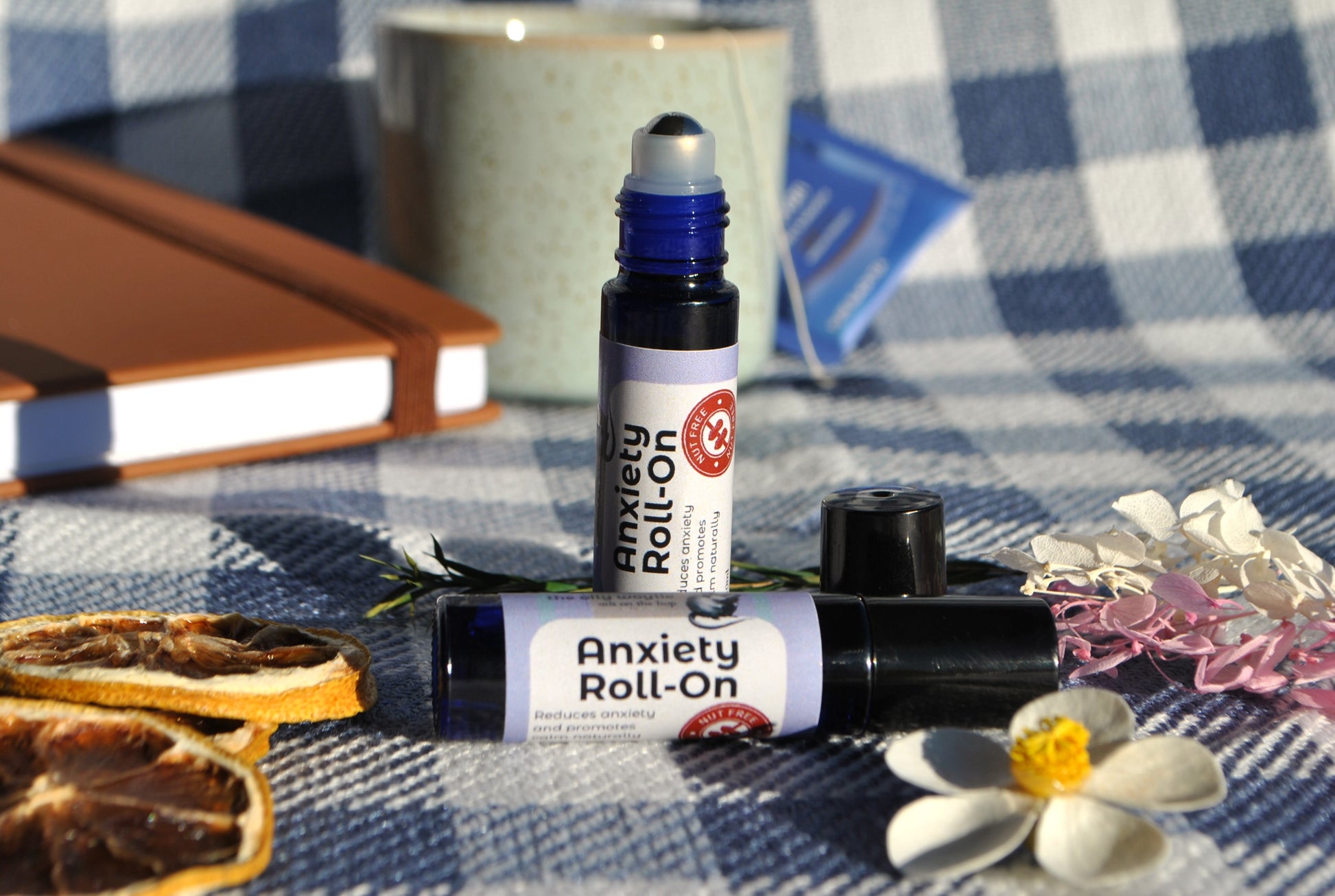 Anxiety Roll essential oil roller blend, natural calming support for stress and anxiety on a checkered tablecloth with flowers and dried oranges.
