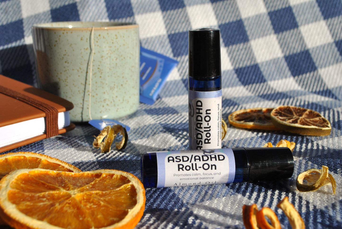 ASD/ADHD Roll essential oil blend in roller bottle, natural calming support on a checkered tablecloth with oranges and a notebook.