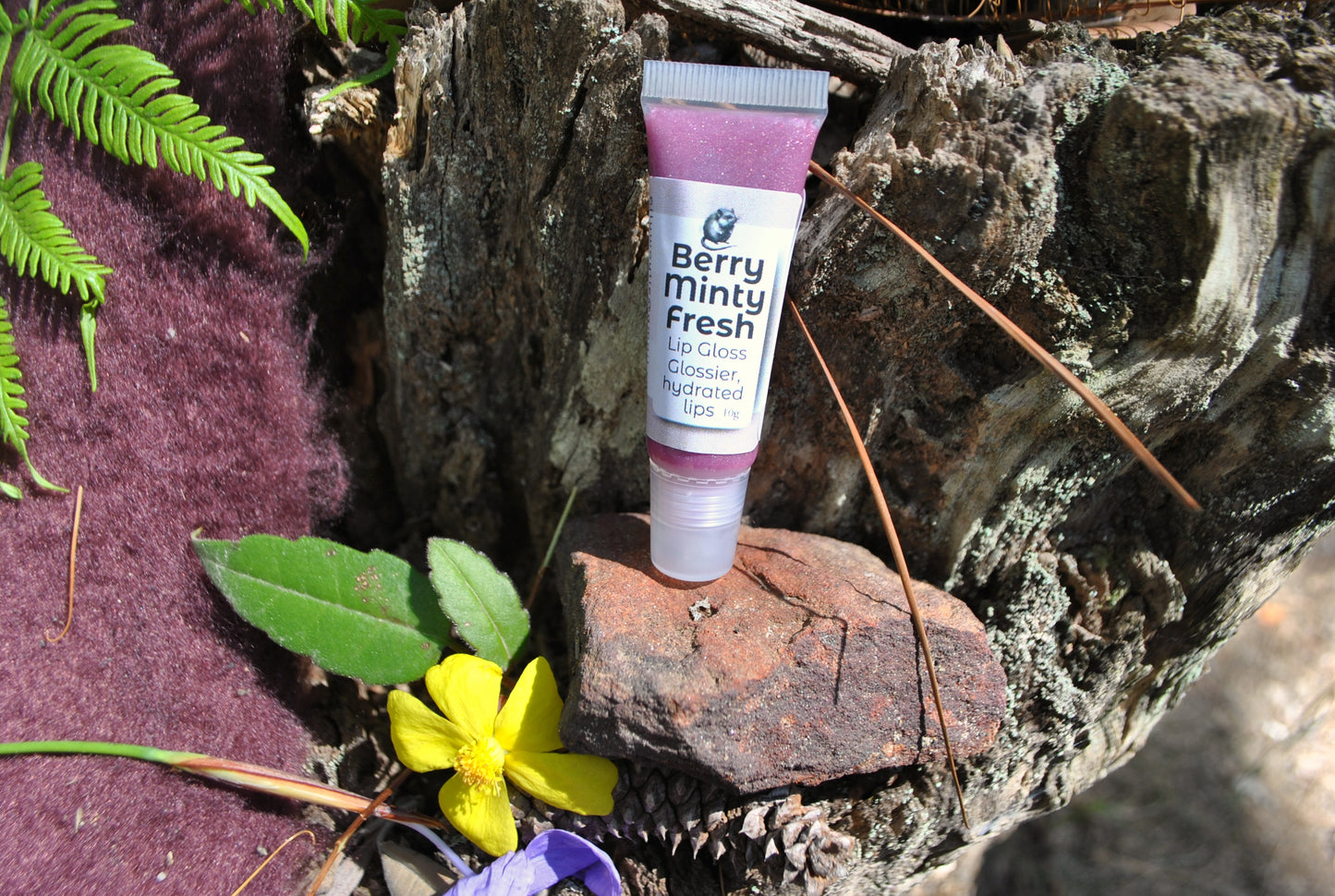 Berry Minty Fresh Lip Gloss by The Oily Woylie resting on rustic bark with fern and yellow wildflower — natural glossy hydration with a cool minty berry shine.