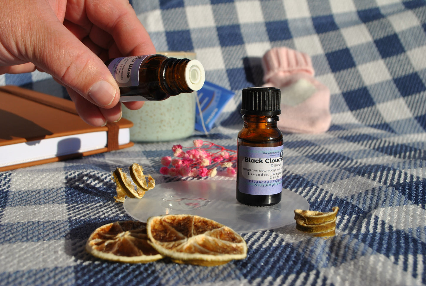 Black Clouds Diffuser essential oil blend bottle, natural support for low mood Hand holding a bottle over a checkered tablecloth with dried oranges and flowers.