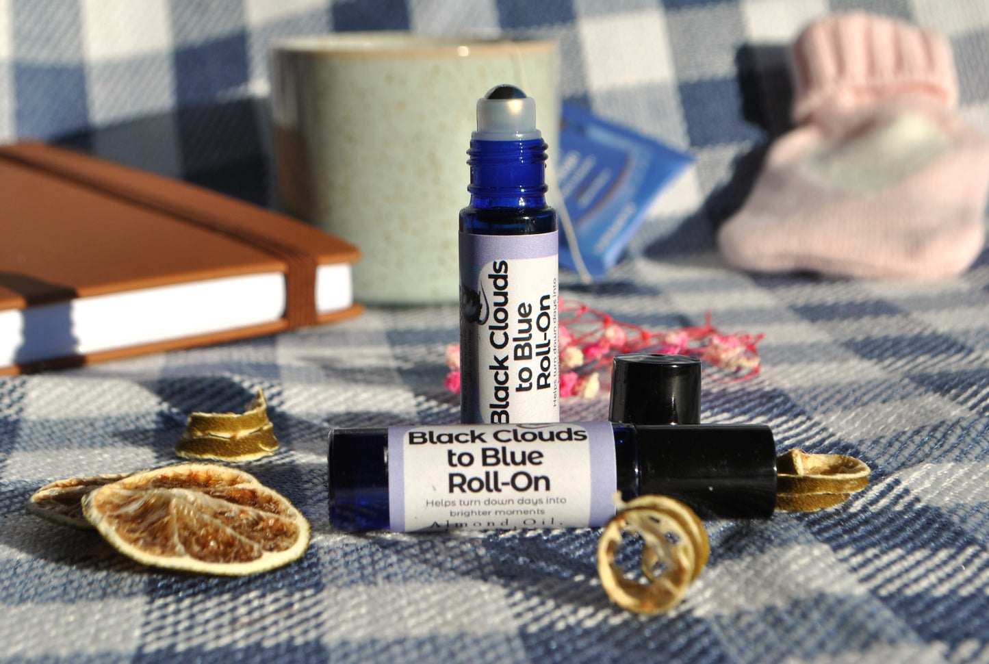 Black Clouds Roll essential oil roller blend, natural support for low mood on a checkered fabric background with dried leaves and a notebook. Lid off