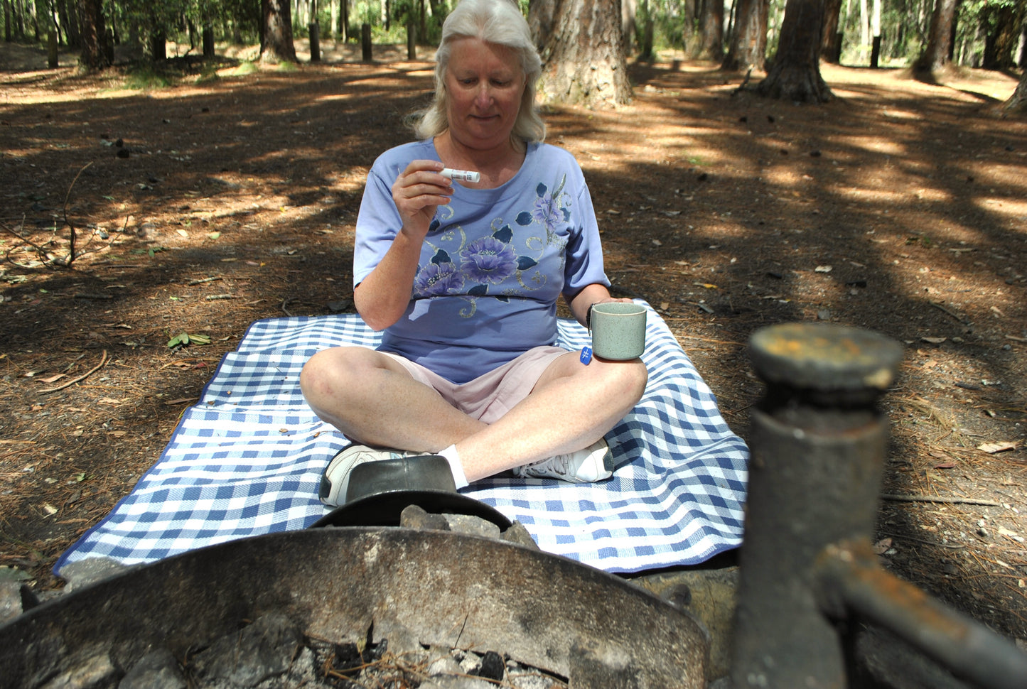 Person sitting cross-legged on a blue check picnic blanket in the Australian bush, holding The Oily Woylie Bug Bite Relief Stick near a campfire — natural remedy for soothing itchy bites.