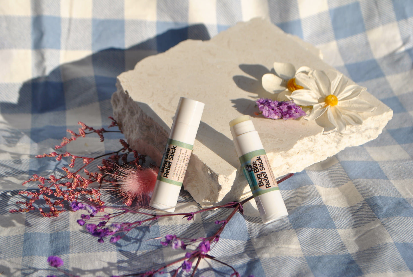 Bug Bite Relief Stick tube, natural soothing balm for insect bites on a stone surface with flowers and a checkered fabric background