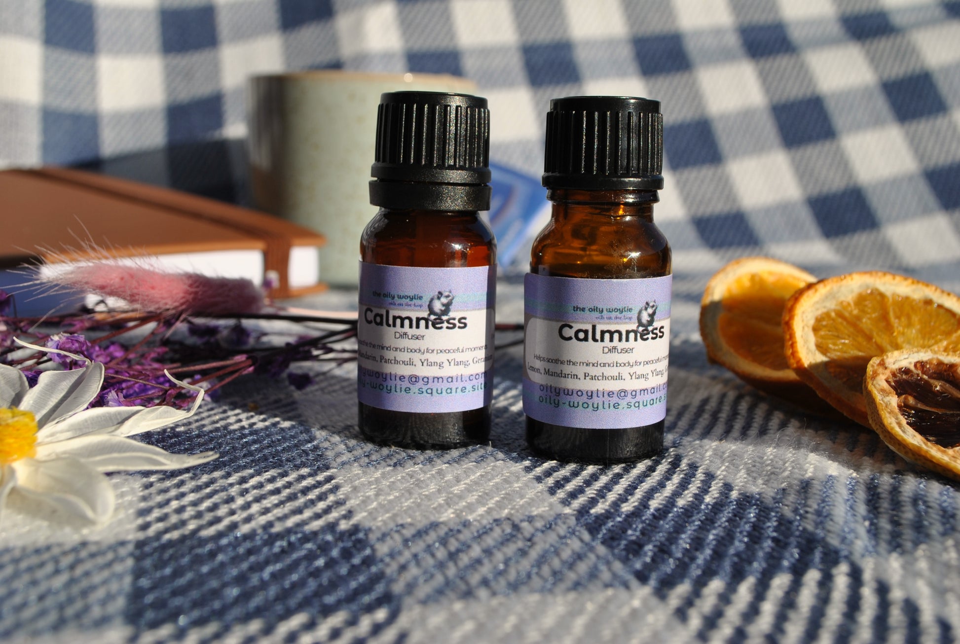 Calm Diffuser essential oil blend bottle, promotes relaxation and reduces stress Two bottles on a checkered tablecloth with dried oranges and flowers.