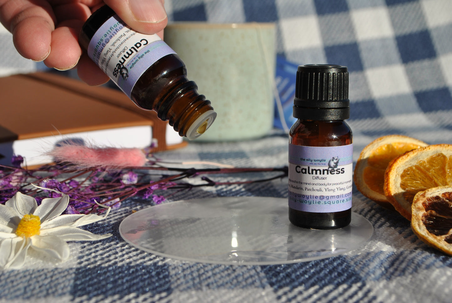 Calm Diffuser essential oil blend bottle, promotes relaxation and reduces stress dripping on a checkered tablecloth with dried oranges and flowers.