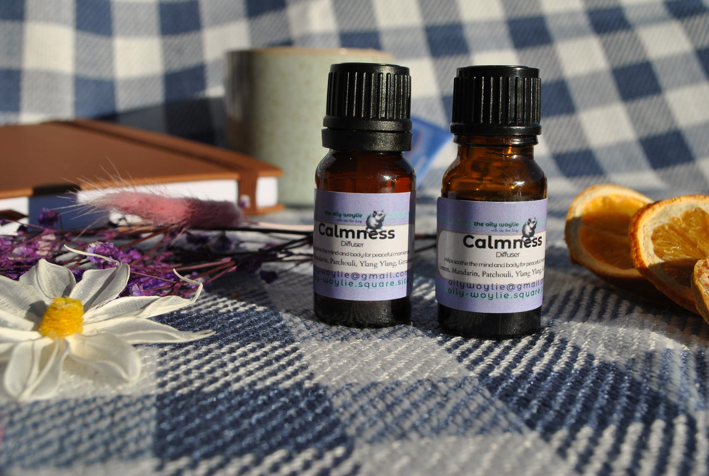 Calm Diffuser essential oil blend bottle, promotes relaxation and reduces stress on a checkered fabric background with flowers and oranges.