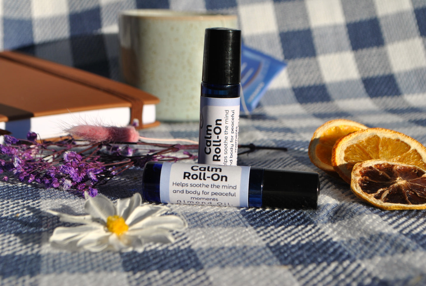 Calm Roll essential oil roller blend, promotes relaxation and stress relief on a checkered tablecloth with flowers and oranges