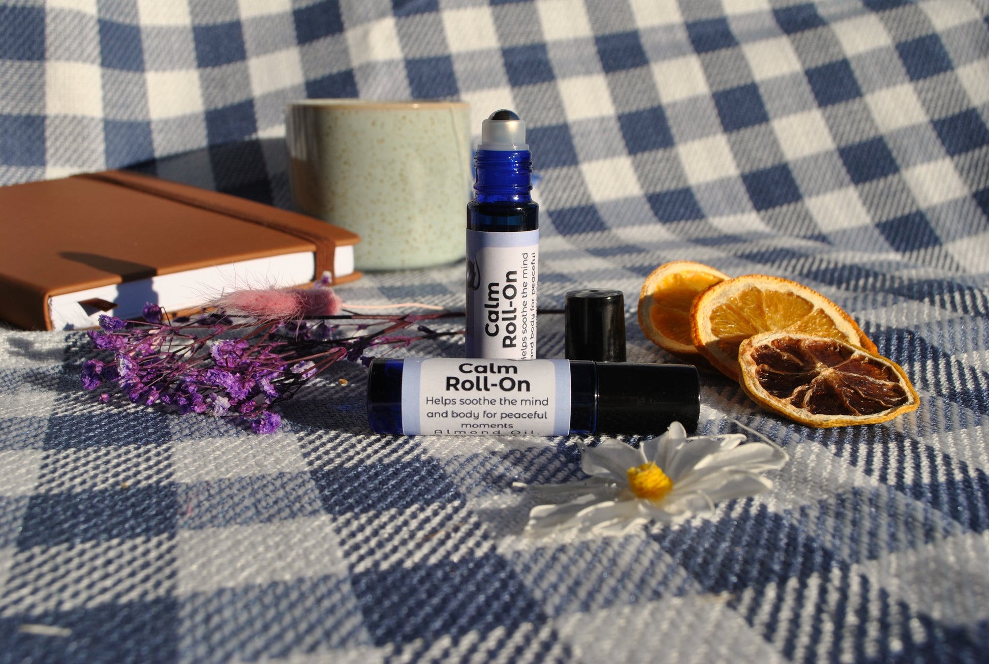 Calm Roll essential oil roller blend, promotes relaxation and stress relief on a checkered fabric background with dried oranges, lavender, and a notebook. Lid off