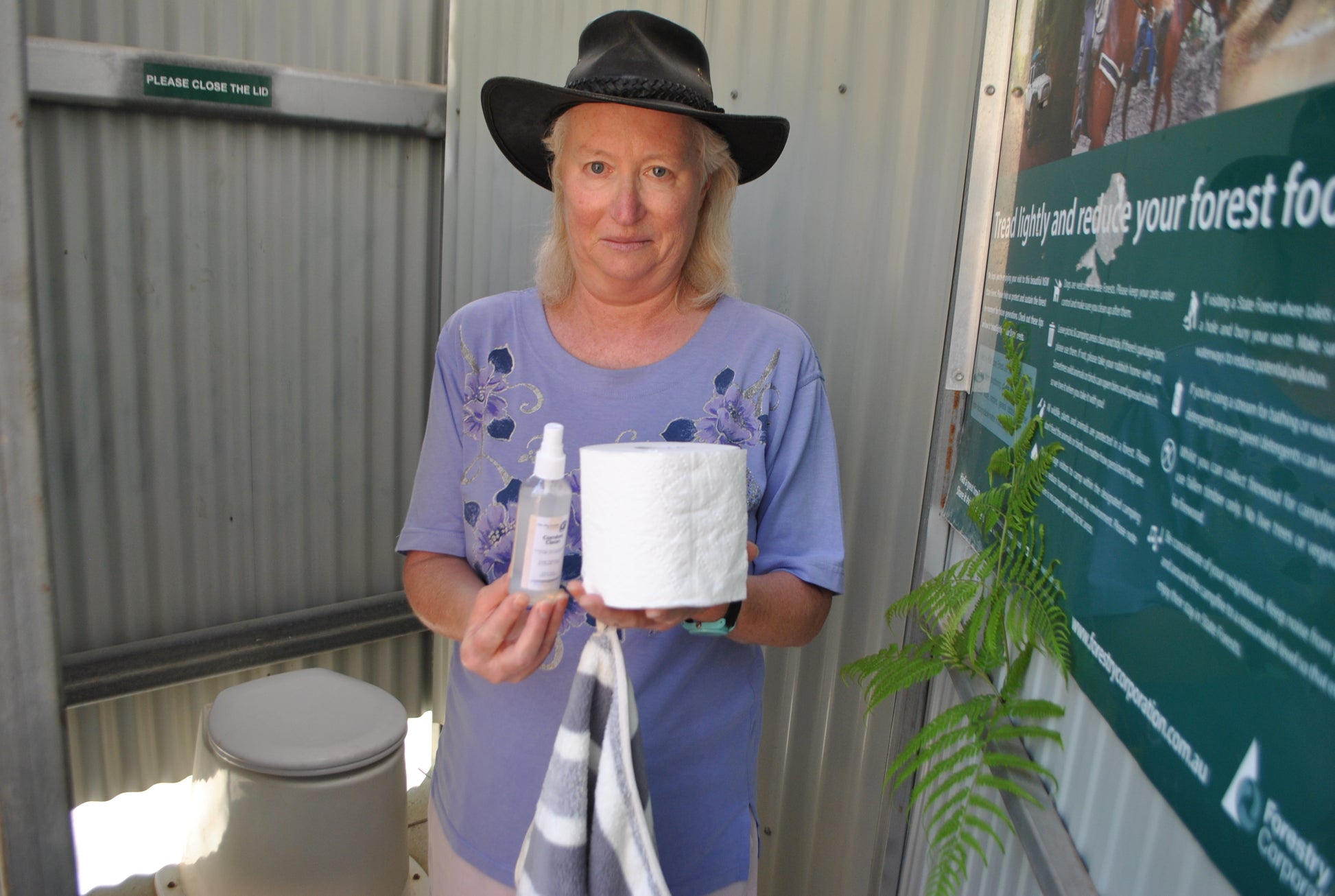 Person holding The Oily Woylie Comfort Clean Spray and a toilet roll in an outdoor bathroom, demonstrating natural hygiene with fern greenery and eco-friendly setting.