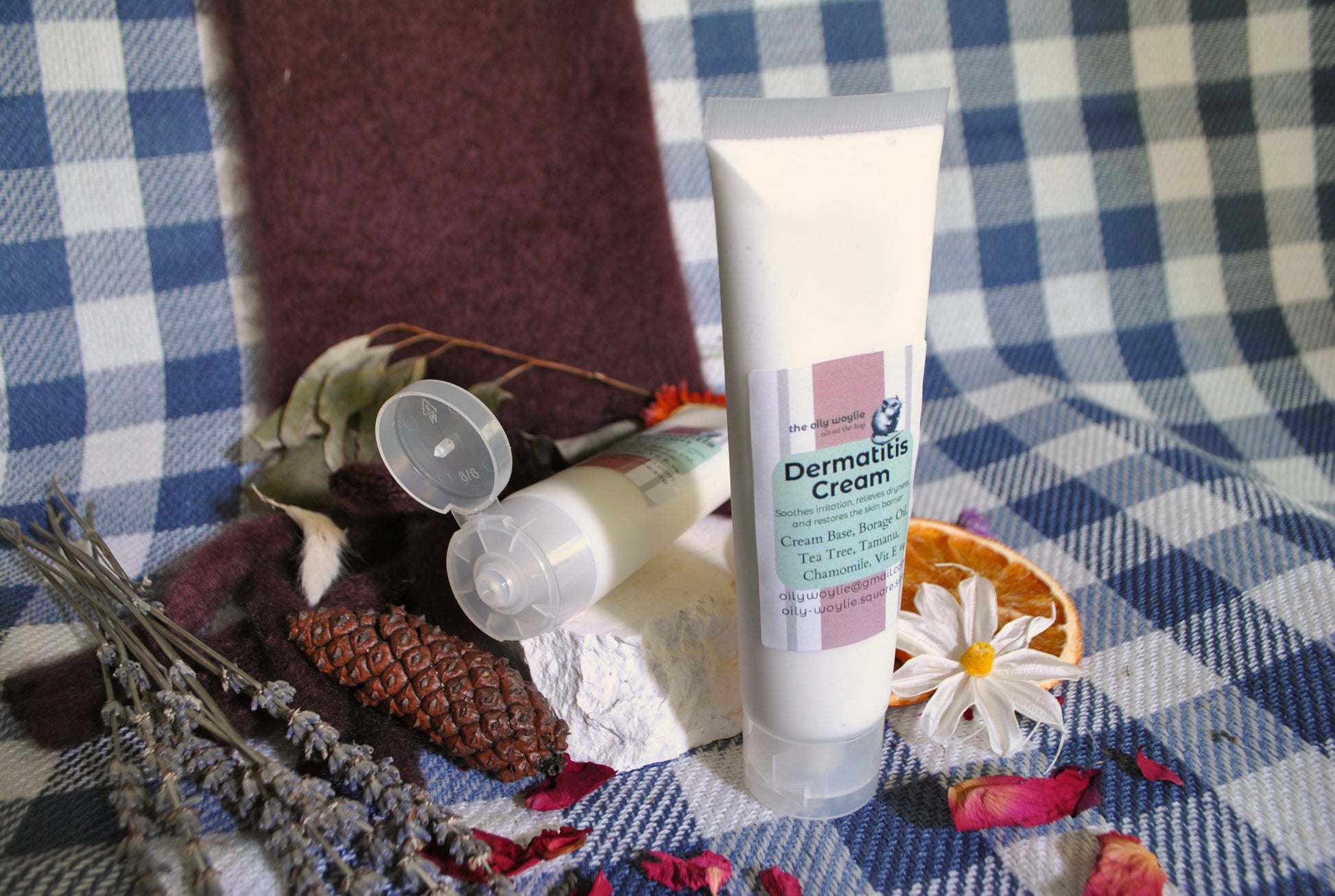 Dermatitis Cream tube, natural cream to soothe irritated and sensitive skin on a checkered fabric background with flowers and herbs.