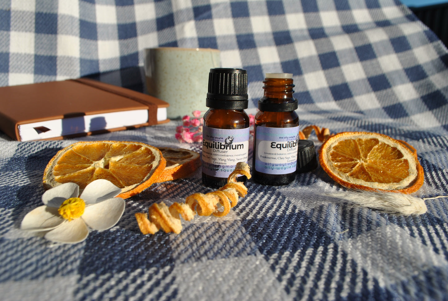Equilibrium Diffuser essential oil blend bottle, promotes balance and calm Two essential oil bottles on a checkered fabric background with dried oranges and flowers.