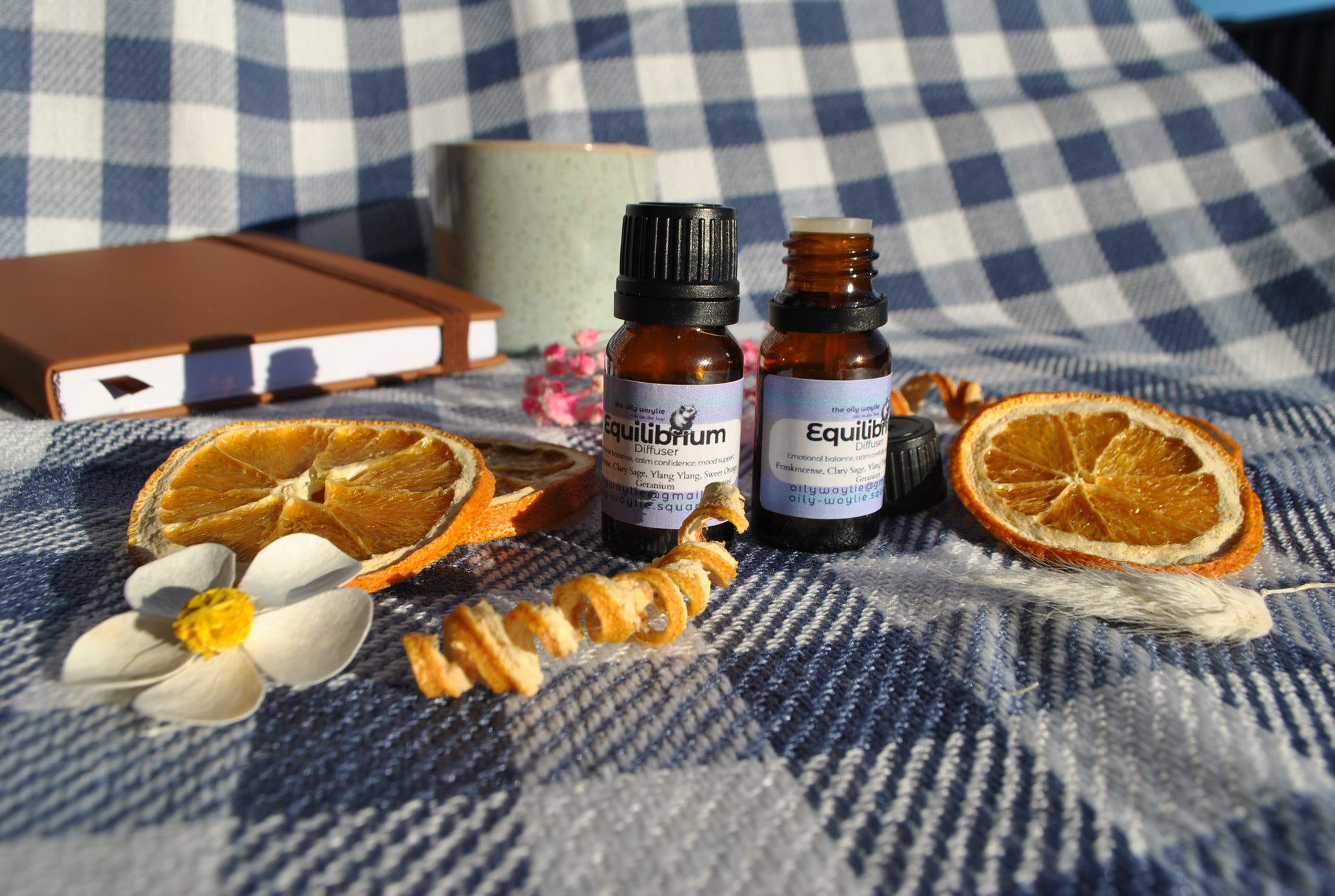 Equilibrium Diffuser essential oil blend bottle, promotes balance and calm Two essential oil bottles on a checkered fabric background with dried oranges and flowers.