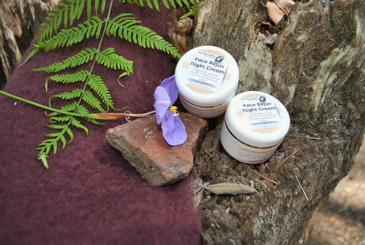 Two jars of Face Rejuv Night Cream resting on a rustic tree stump with fern leaves and a purple wildflower — soothing natural bush skincare by The Oily Woylie.