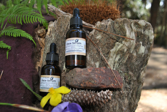 The Oily Woylie Face Serum bottles resting on rustic bark with fern and wattle flowers — natural hydrating serum with essential oils to nourish and restore radiant skin.