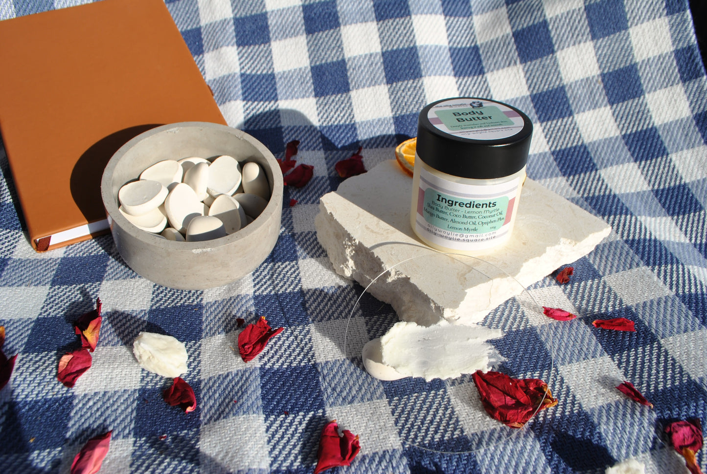 Lemon Myrtle Body Butter jar, natural moisturising body cream with lemon myrtle scent and bowl of white stones on a checkered tablecloth with flowers.