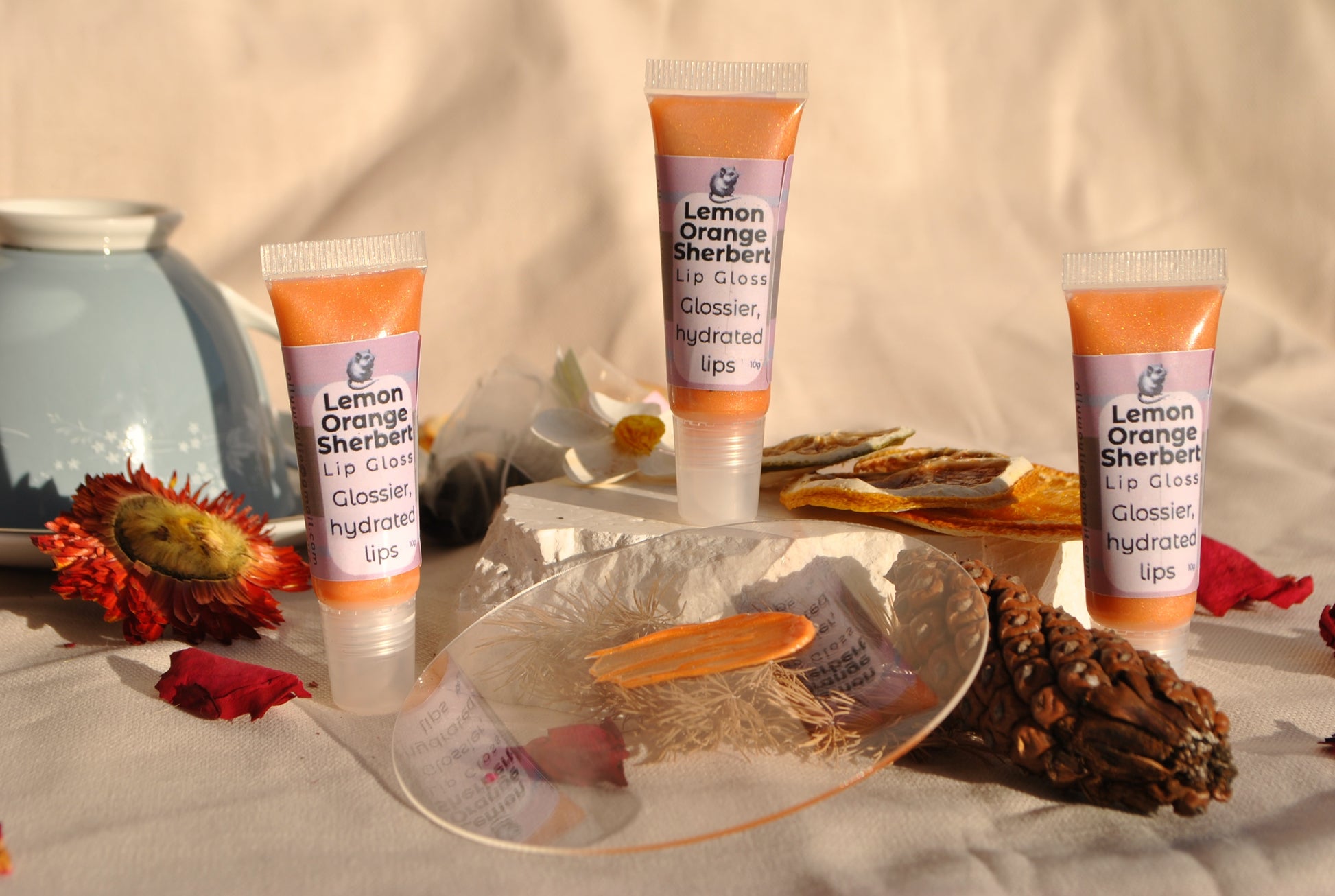 Lemon Orange Sherbet lip gloss tube, natural glossy lip colour with citrus sherbet scent on a decorative surface with flowers and a bowl.