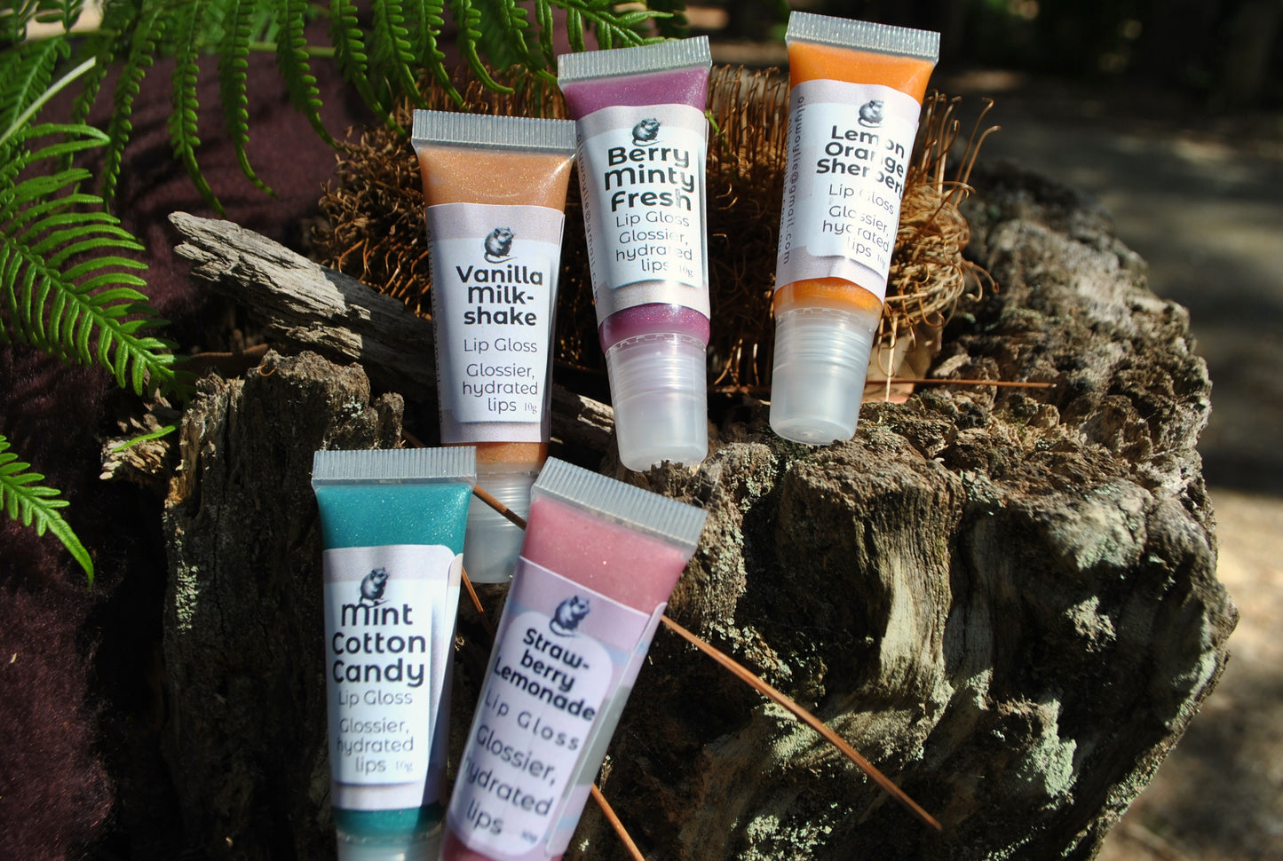 Five tubes of natural lip gloss by The Oily Woylie — Vanilla Milkshake, Berry Minty Fresh, Lemon Orange Sherbet, Strawberry Lemonade, and Mint Cotton Candy — displayed on rustic bark with ferns and sunlight.