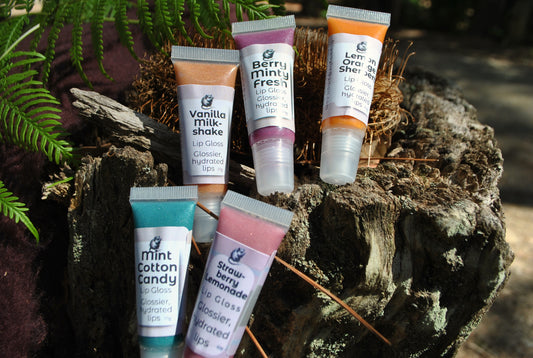 Five tubes of natural lip gloss by The Oily Woylie — Vanilla Milkshake, Berry Minty Fresh, Lemon Orange Sherbet, Strawberry Lemonade, and Mint Cotton Candy — displayed on rustic bark with ferns and sunlight.