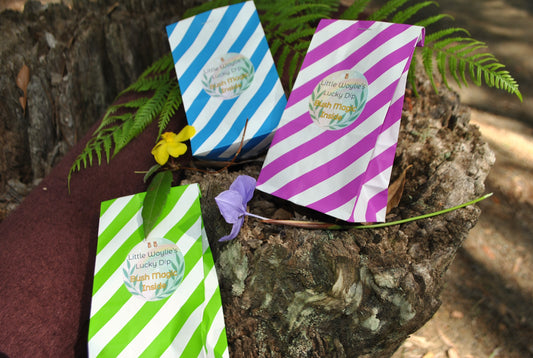 Three striped Little Woylie’s Lucky Dip bags in green, blue, and purple, resting on a tree stump with ferns and bush flowers — playful Aussie bush setting by The Oily Woylie.