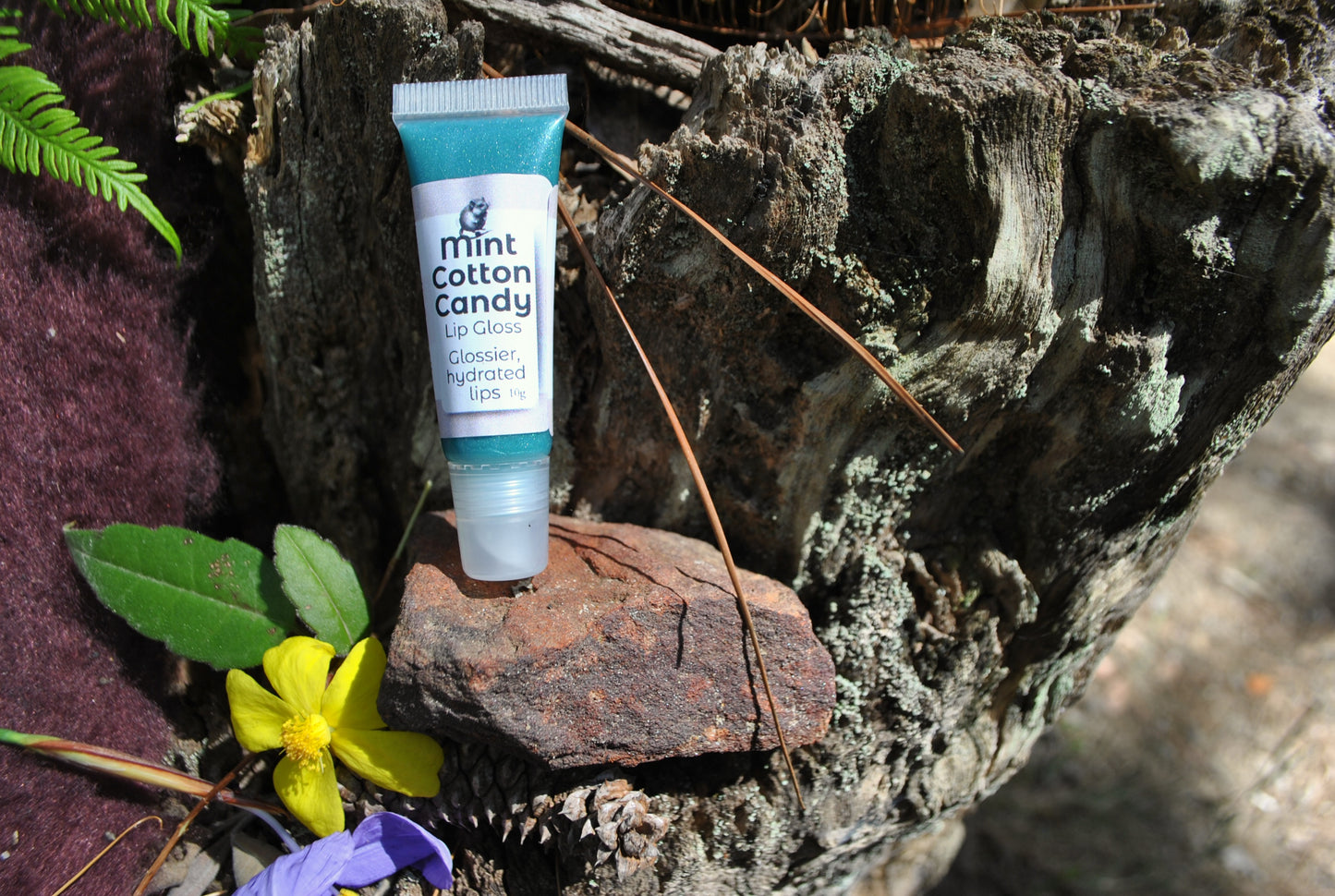 Mint Cotton Candy Lip Gloss by The Oily Woylie resting on rustic bark with yellow and purple wildflowers — natural, glossy hydration for lips with a sweet minty twist.