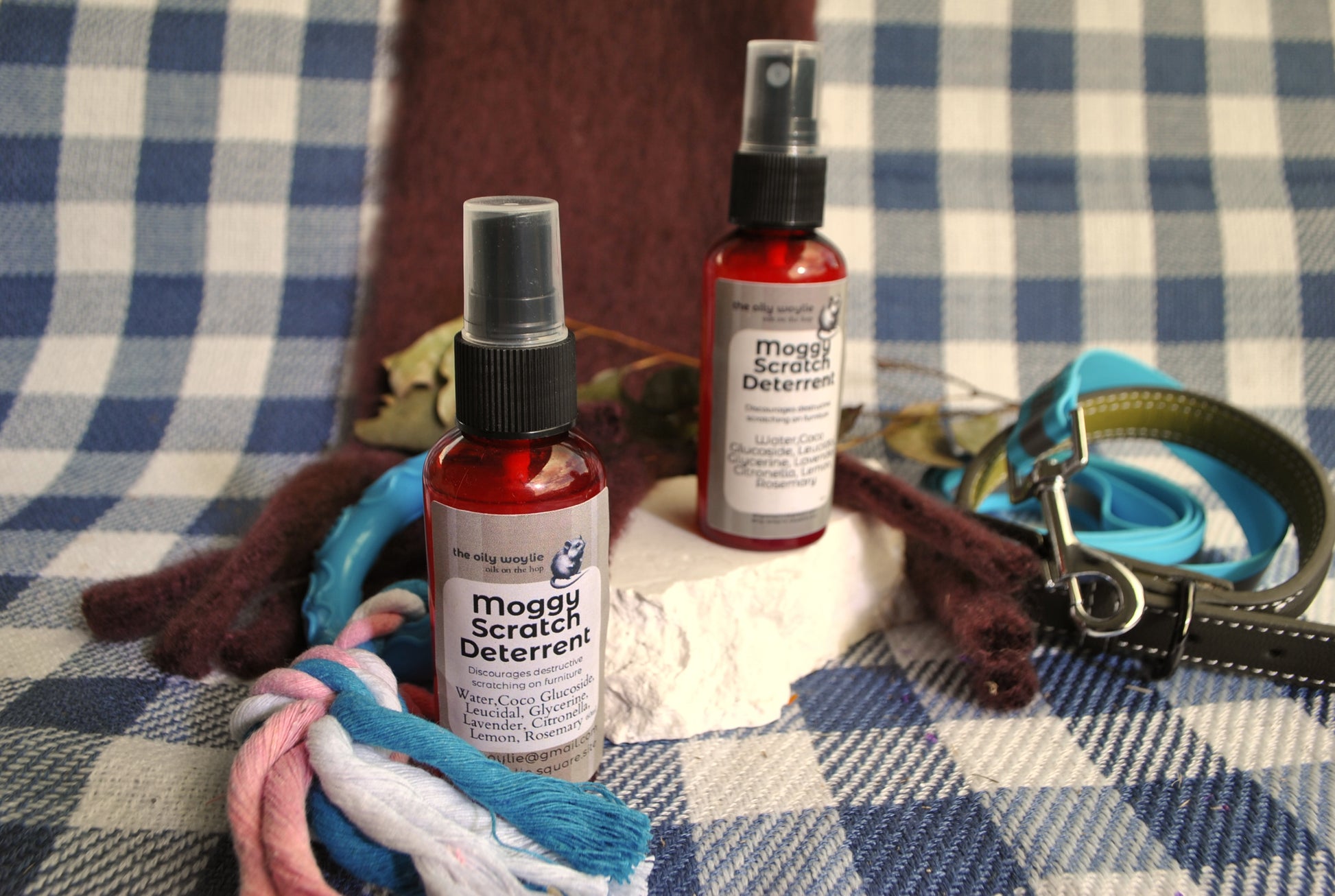 Moggy Scratch Deterrent spray bottle, natural spray to discourage cats from scratching furniture on a checkered fabric background with a dog toy and leash.