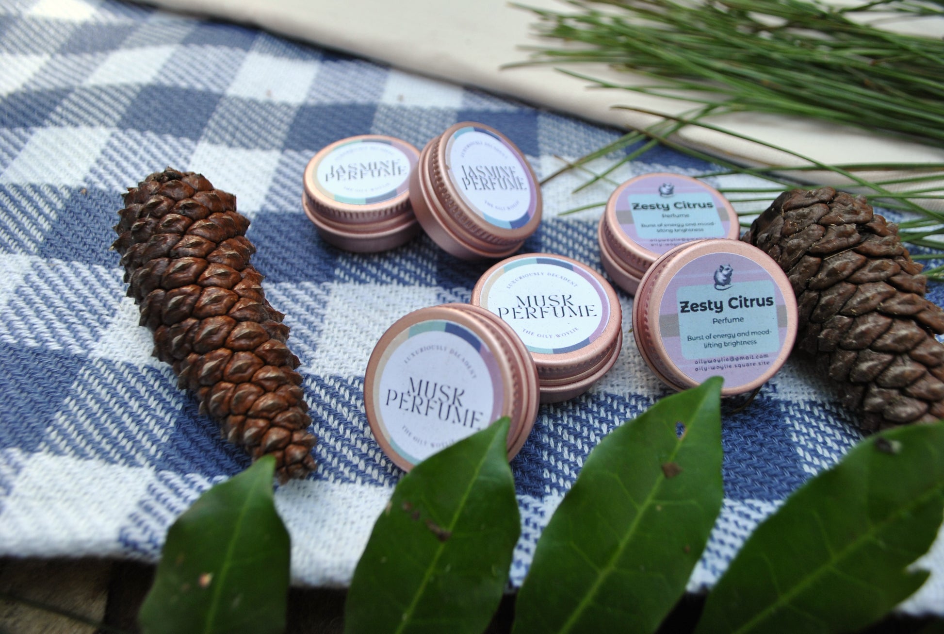 The Oily Woylie natural perfume range, Jasmine, Musk, and Zesty Citrus scents, handmade with essential oils on a checkered cloth with pine cones and leaves.