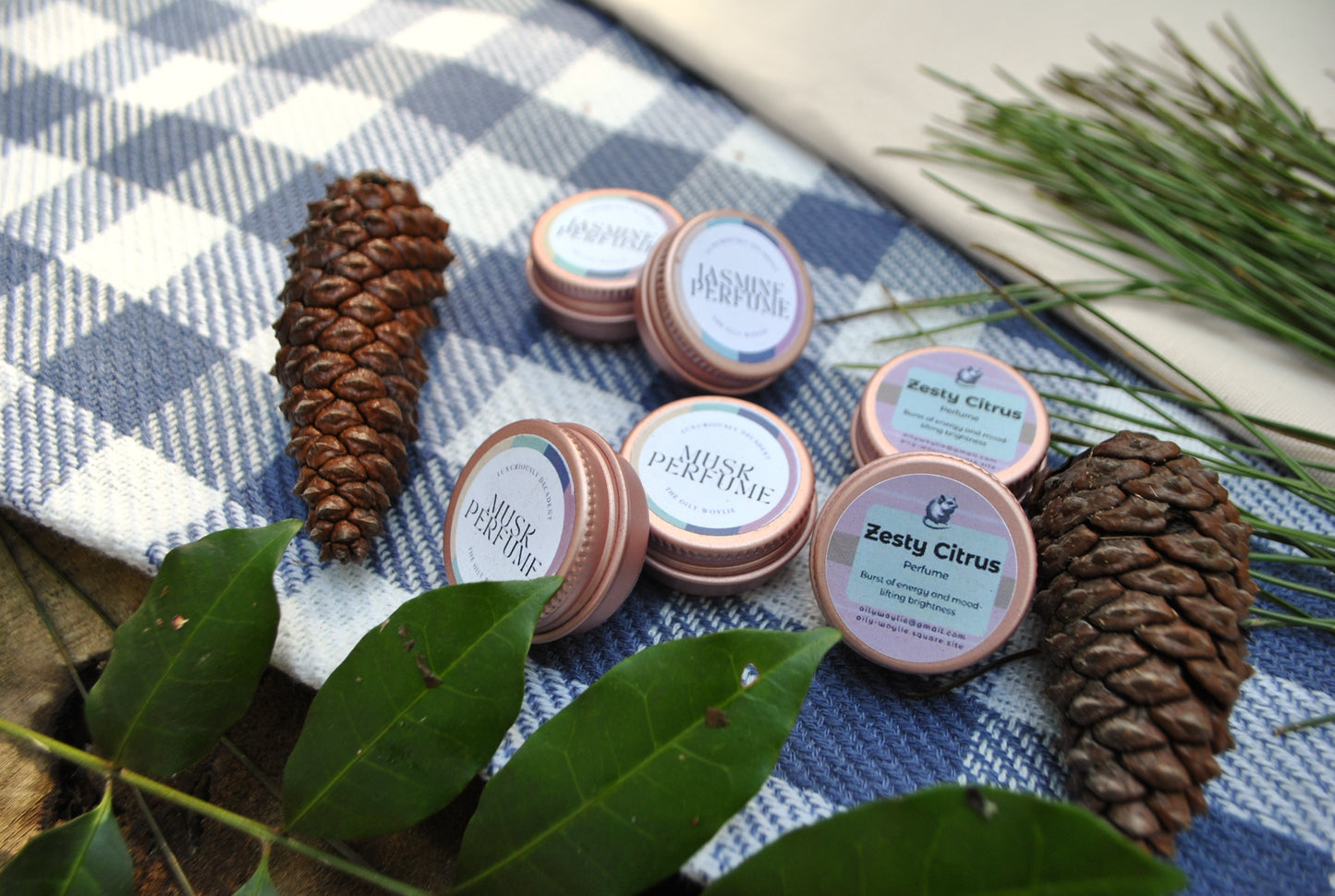 The Oily Woylie natural perfume range, Jasmine, Musk, and Zesty Citrus scents, handmade with essential oils and Pine on a checkered cloth with pine cones and leaves.