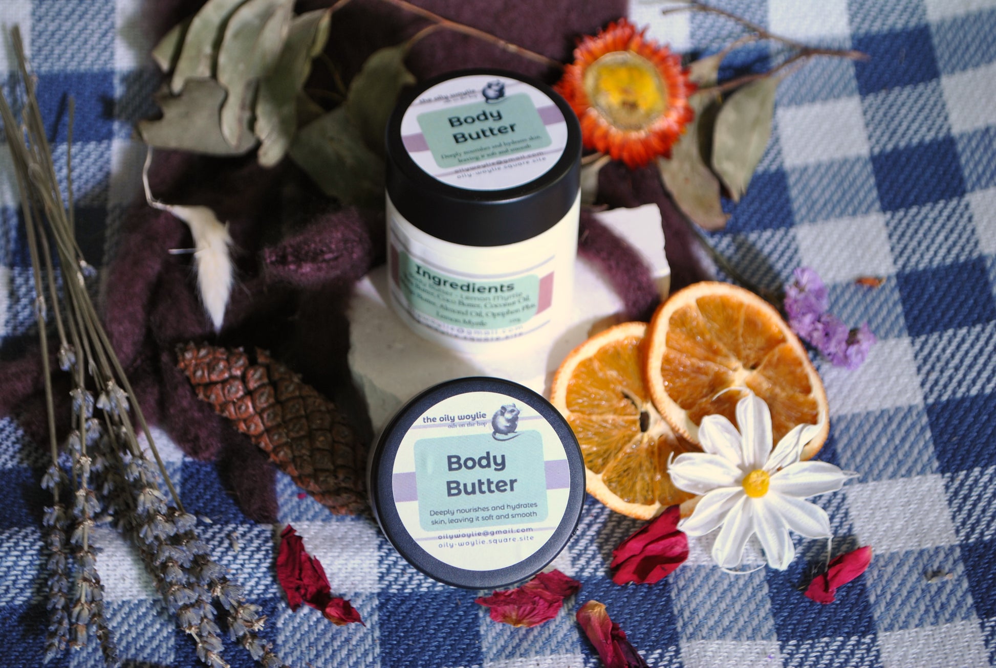 Rose and Lemon Myrtle Body Butter jars side by side, natural moisturising body creams on a checkered fabric background with dried oranges and flowers.