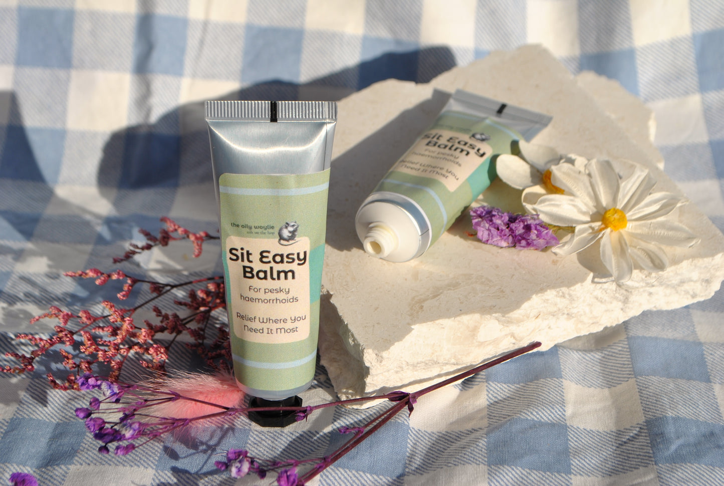 Sit Easy Balm tube, natural soothing balm for haemorrhoid discomfort on a checkered fabric background with flowers.