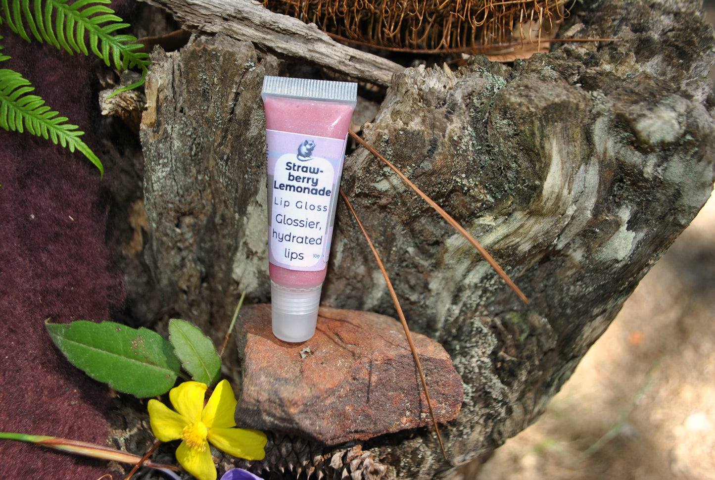 Strawberry Lemonade Lip Gloss by The Oily Woylie resting on rustic bark with fern and yellow wildflower — sweet pink shine with natural hydration for glossy lips.