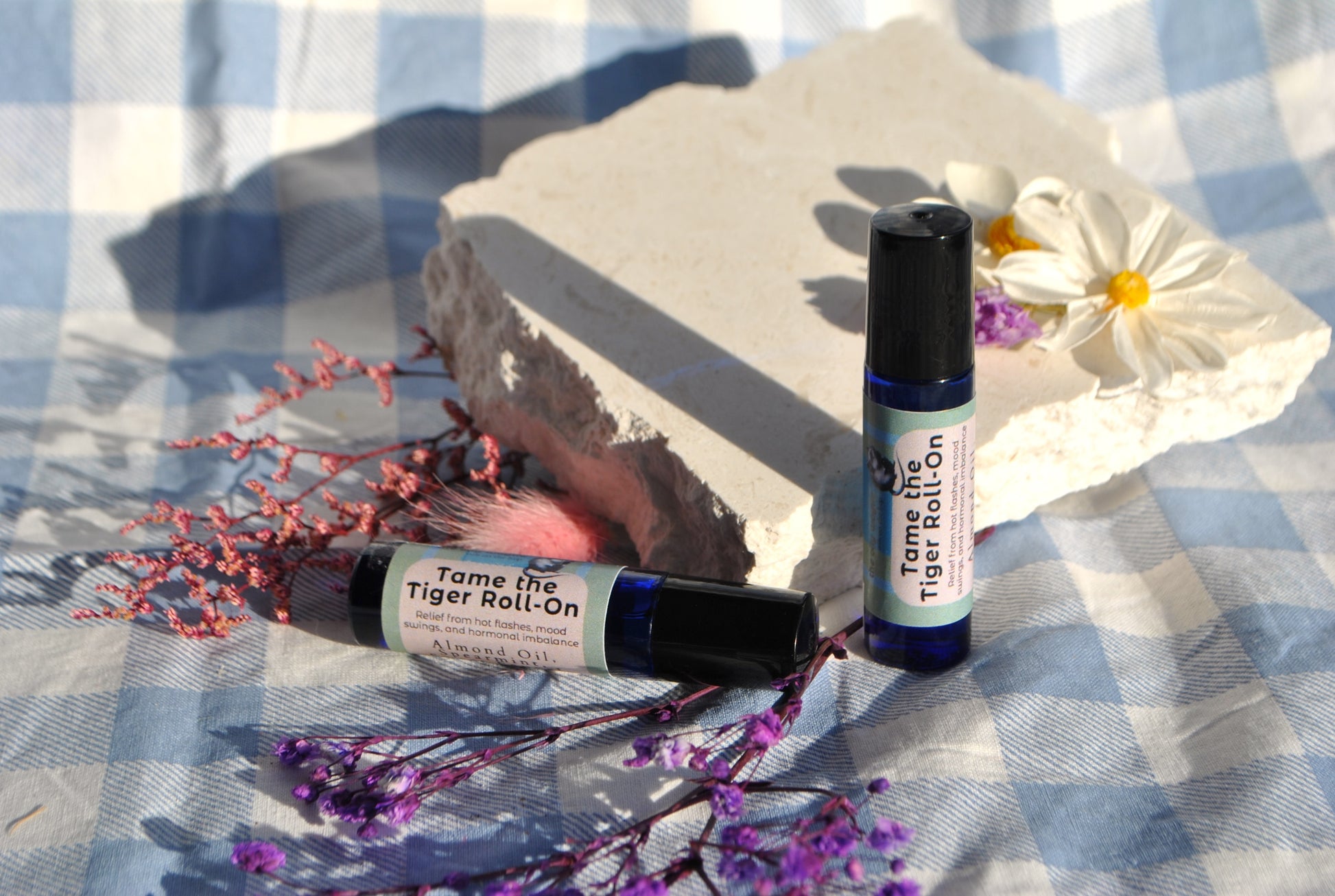 Tame the Tiger Roll essential oil roller blend, natural support for hormones and mood on a checkered fabric background with flowers and rocks.