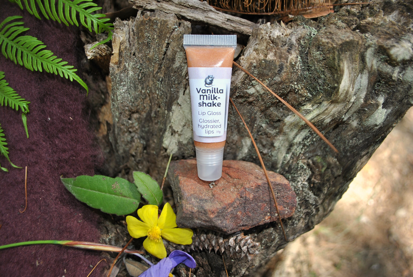 Vanilla Milkshake Lip Gloss by The Oily Woylie resting on rustic bark with fern and yellow wildflower — creamy golden gloss for soft, hydrated, naturally glossy lips.