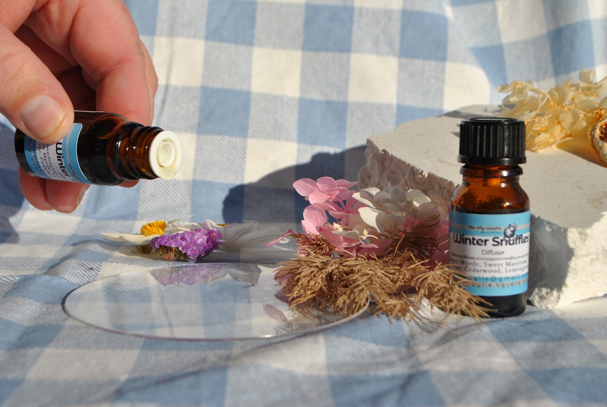 Winter Snuffles Diffuser Blend essential oil bottle, natural support for colds and congestion onto flowers on a checkered tablecloth.