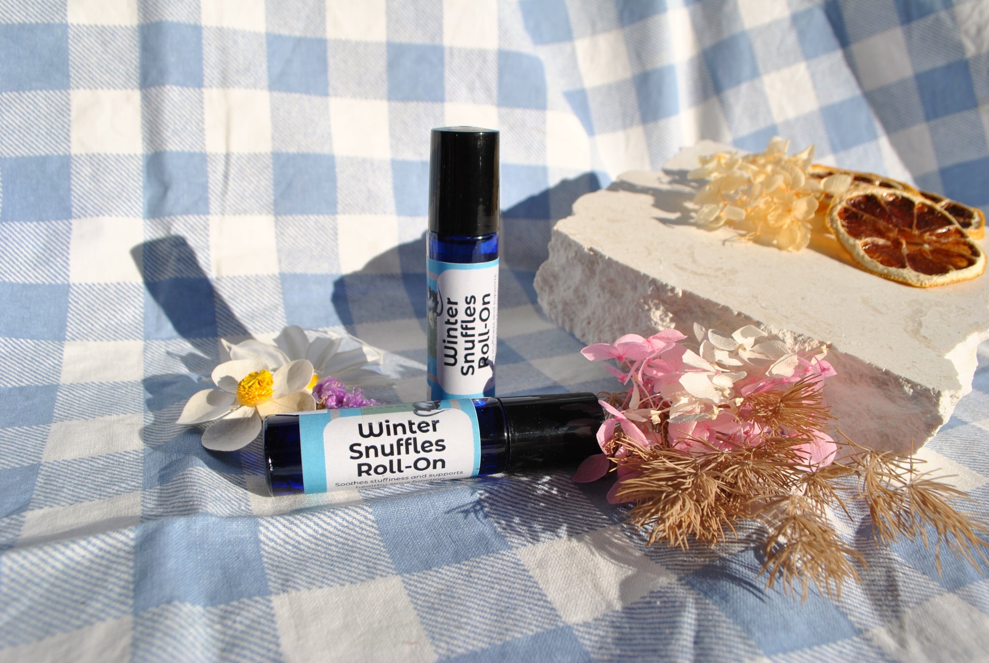 Winter Snuffles Roll essential oil roller blend, natural support for colds and congestion on a blue and white checkered fabric with flowers and dried plants.