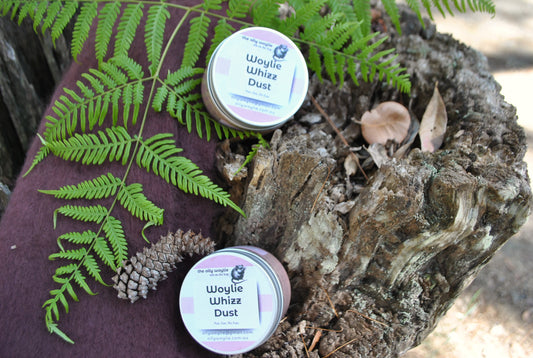 Two tins of Woylie Whizz Dust bath fizz resting on a rustic tree stump with green fern leaves and pine cone — natural bush setting by The Oily Woylie.
